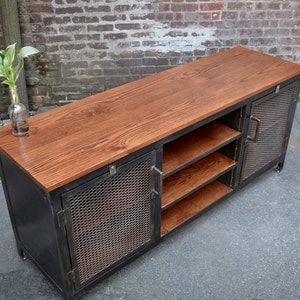 May include: A rustic, industrial-style media console. It features a dark metal frame with mesh door panels and a warm-toned wooden top and shelves. The console has a rectangular shape and is designed for storage and display.