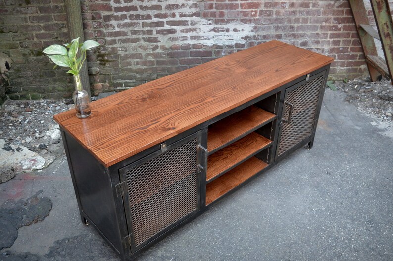 May include: A rustic, industrial-style media console. It features a dark metal frame with mesh door panels and a warm-toned wooden top and shelves. The console has a rectangular shape and is designed for storage and display.