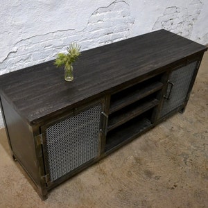 May include: A dark-stained wooden media console with metal mesh doors and a matching top. The console has open shelving and a small glass vase with greenery on top. The industrial-style cabinet is suitable for a living room.
