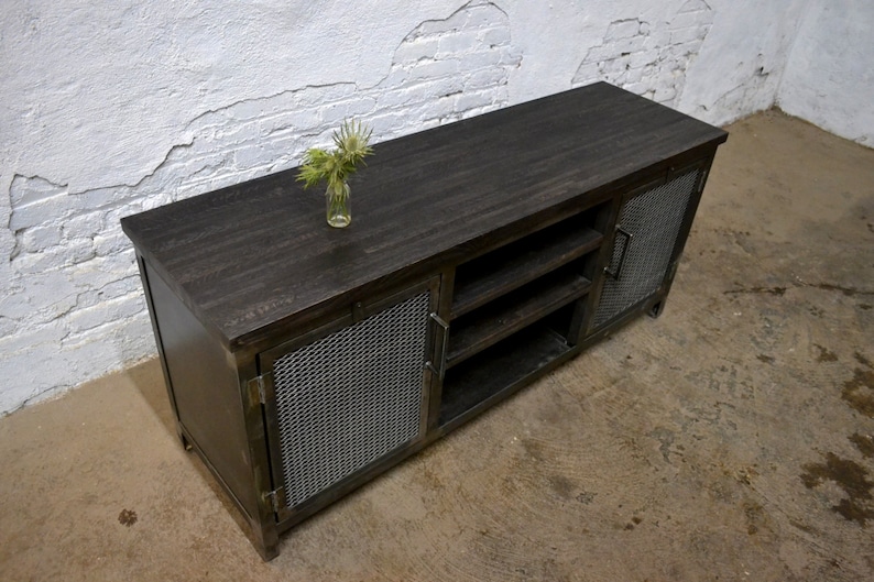 May include: A dark-stained wooden media console with metal mesh doors and a matching top. The console has open shelving and a small glass vase with greenery on top. The industrial-style cabinet is suitable for a living room.