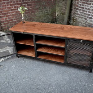 May include: A rustic, industrial-style cabinet with a reddish-brown wooden top and shelves. The cabinet has a black metal frame and a door with a mesh design. The cabinet is designed for storage.