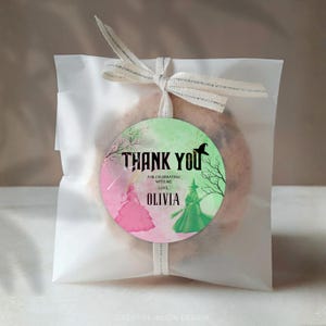 May include: A cookie in a clear cellophane bag tied with a ribbon. A round tag with the words "THANK YOU" and "FOR CELEBRATING WITH ME, LOVE, OLIVIA" is attached. The tag features a pink and green watercolor design with witch and princess illustrations.