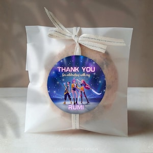 May include: A clear cellophane bag tied with a cream ribbon, holding a cookie with a round sticker. The sticker reads "THANK YOU for celebrating with me" and features three animated figures. The name "RUMI" is printed below.