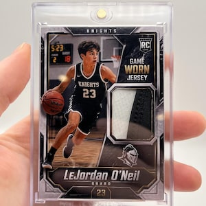 Op de afbeelding: Een basketbalkaart van LeJordan O'Neil in een beschermhoesje. De kaart toont een speler in een zwart-wit Knights-shirt, nummer 23. Het bevat een stukje wedstrijdshirt en is gelabeld met "Rookie Card".