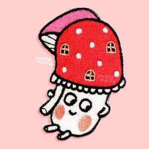 May include: An embroidered patch featuring a white mushroom character with a red cap decorated with white spots and small arched windows. The character has a smiling face with pink cheeks, black eyes, and small arms and legs. The text Missy Minzy is visible.