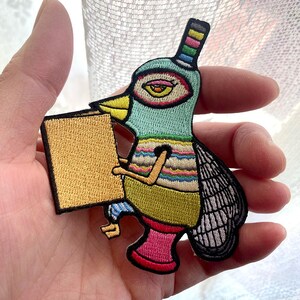 May include: Embroidered patch of a quirky bird holding a golden rectangle. The bird has a teal head, yellow beak, and a striped body with pink boots. The patch has a black border and is held in a hand.