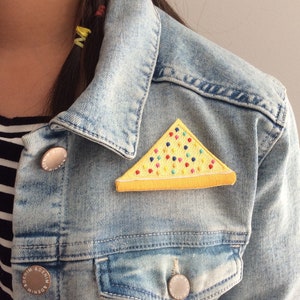 May include: A yellow triangle-shaped patch with colourful sprinkles on a denim jacket. The patch is a representation of a piece of Australian food called 'Fairy Bread'.