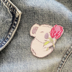 May include: Embroidered patch of a light pink koala holding a pink flower with green leaves. The koala has purple accents. The patch is sewn onto a denim jacket.