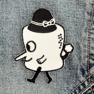 May include: A white embroidered patch featuring a cartoon character with a long nose, one large eye, and a black bowler hat with a white bow. The character has black limbs and a small white tail, shown against a blue denim fabric background.