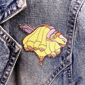 May include: A yellow and purple embroidered patch of a hot air balloon with a basket and a purple tail. The patch is sewn onto a blue denim jacket.