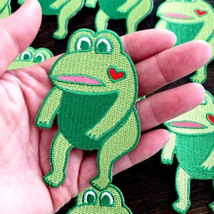 May include: Embroidered green frog patches with a heart detail. The patches feature a light green body, dark green belly, and pink mouth. The frog patches are a fun craft supply.