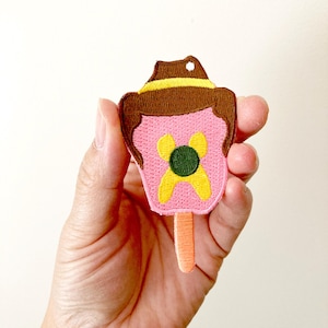 May include: An embroidered iron-on patch of a pink popsicle with a brown hat and a yellow flower with a green centre. The popsicle has a stick.