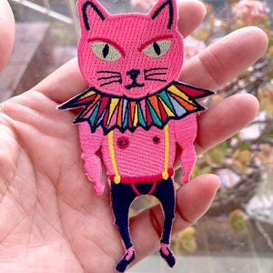 May include: A pink embroidered cat patch with a stern expression. The cat has green eyes, a colourful collar, and is wearing blue trousers with yellow braces. The patch is held in a hand, with a blurred background of greenery.