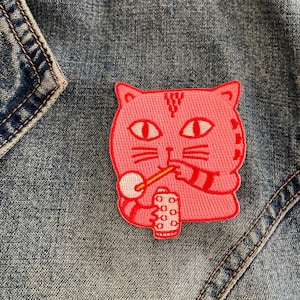May include: Pink embroidered iron-on patch of a cat drinking from a can with a straw. The cat has a pink body, white eyes, and black whiskers.