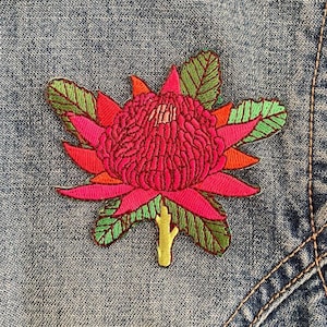 May include: An embroidered patch featuring a vibrant pink and red waratah flower with textured petals, surrounded by green leaves and a small yellow stem, stitched onto a blue denim fabric background with visible orange thread seams.
