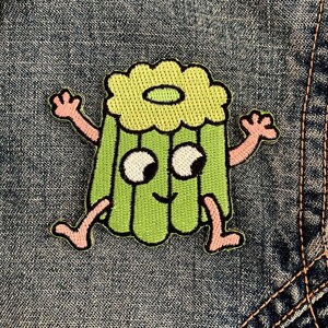 May include: An embroidered patch featuring a light green gelatin dessert character with a ruffled top, two large white eyes with black pupils, a small smile, and thin pink arms and legs extended outward, outlined in black thread.