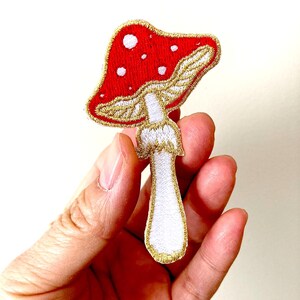 May include: An embroidered mushroom patch in red and white with gold accents. The mushroom has white spots on the red cap and a white stem with gold outlining.