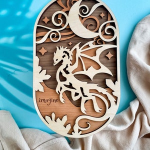 May include: A wooden wall hanging with a crescent moon, stars, and a dragon in a forest setting. The words "imagine" are engraved on the bottom of the oval-shaped piece.