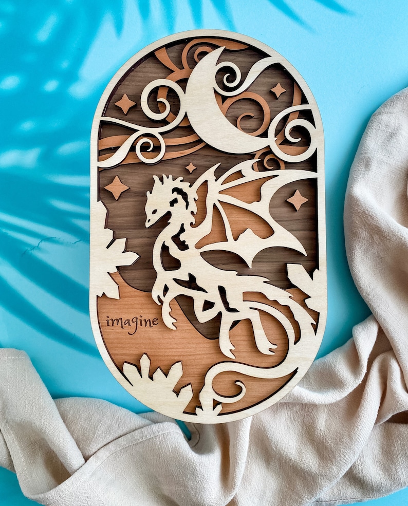 May include: A wooden wall hanging with a crescent moon, stars, and a dragon in a forest setting. The words "imagine" are engraved on the bottom of the oval-shaped piece.