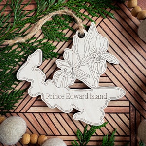 May include: A wooden ornament shaped like Prince Edward Island with a line drawing of a Lady's Slipper orchid. The ornament is on a wooden background.