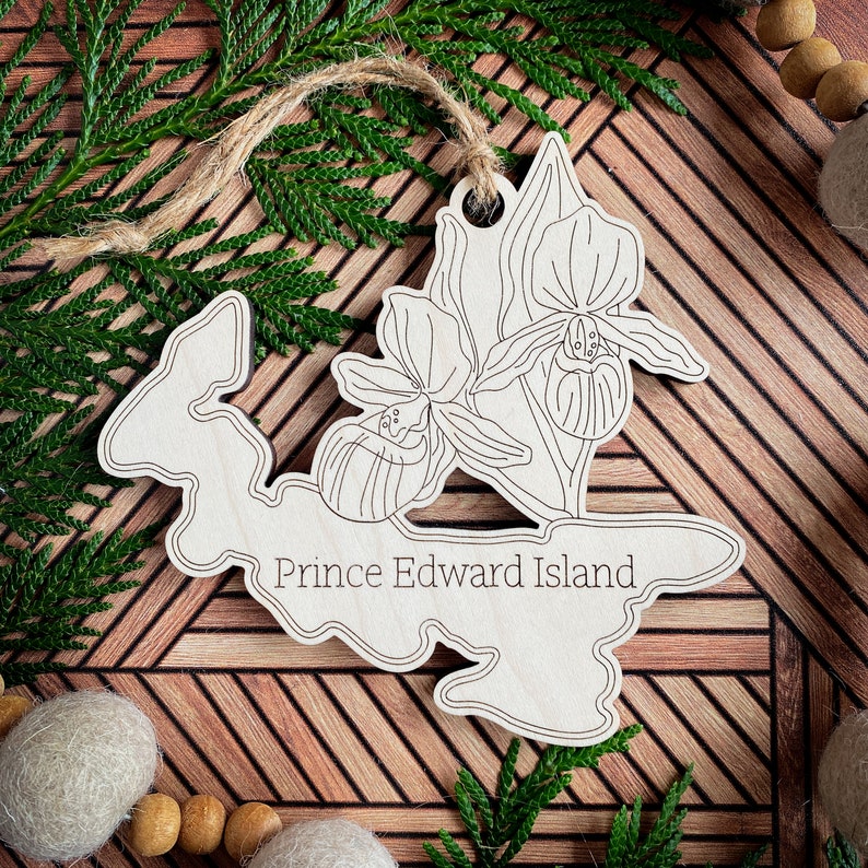 May include: A wooden ornament shaped like Prince Edward Island with a line drawing of a Lady's Slipper orchid. The ornament is on a wooden background.