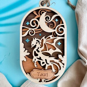 May include: A wooden ornament featuring a dragon silhouette with a crescent moon and stars. The ornament is personalized with the name "Tavi" on a small wooden tag.