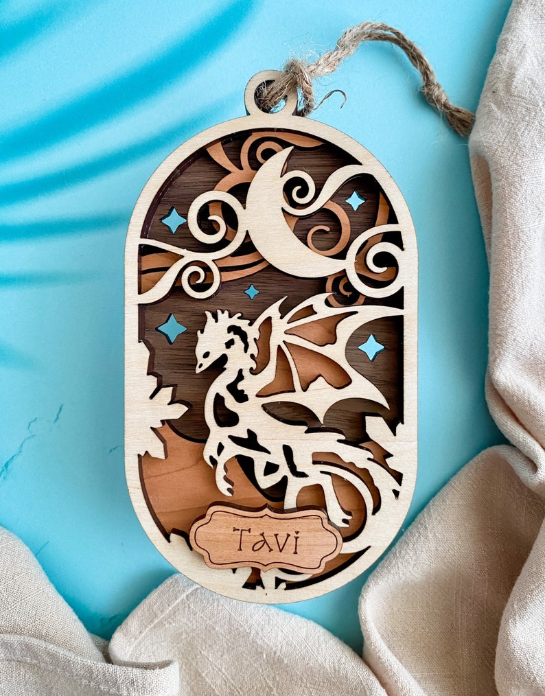 May include: A wooden ornament featuring a dragon silhouette with a crescent moon and stars. The ornament is personalized with the name "Tavi" on a small wooden tag.