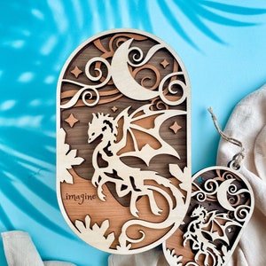 May include: A wooden cutout of a dragon with wings spread, flying in front of a crescent moon and stars. The cutout is painted in shades of brown and has the word "imagine" engraved on the bottom.