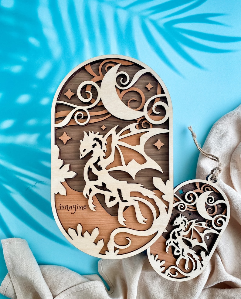 May include: A wooden cutout of a dragon with wings spread, flying in front of a crescent moon and stars. The cutout is painted in shades of brown and has the word "imagine" engraved on the bottom.