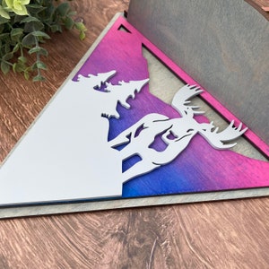 May include: A wooden triangular wall decor with a white moose and trees silhouette against a pink, purple, and blue ombre background.