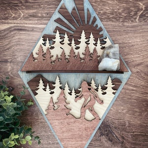 May include: A wooden wall decor in the shape of a diamond with a sunrise scene and a Bigfoot silhouette. The top half of the diamond features a sun with rays and a row of trees. The bottom half features a Bigfoot walking through a forest. The wood is stained in shades of brown and blue.