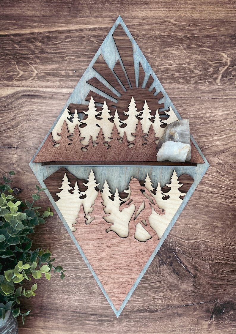 May include: A wooden wall decor in the shape of a diamond with a sunrise scene and a Bigfoot silhouette. The top half of the diamond features a sun with rays and a row of trees. The bottom half features a Bigfoot walking through a forest. The wood is stained in shades of brown and blue.