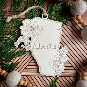 May include: A wooden ornament shaped like the province of Alberta, Canada, with a floral design and the word "Alberta" inscribed on it.