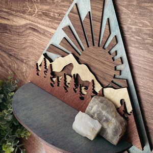May include: A wooden wall shelf shaped like a mountain range with a sun rising behind it. The shelf is painted in shades of blue and brown. Two white and gray crystals are sitting on the shelf.