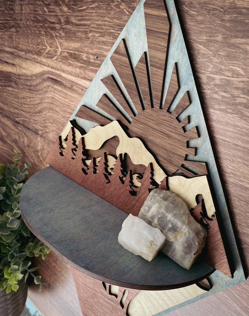May include: A wooden wall shelf shaped like a mountain range with a sun rising behind it. The shelf is painted in shades of blue and brown. Two white and gray crystals are sitting on the shelf.