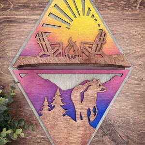 May include: A wooden diamond-shaped wall decor with a split design. The top half features a sunset scene with two chairs and a campfire. The bottom half features a mountain scene with a bear and two trees. The wood is stained in shades of yellow, orange, pink, purple, and blue.