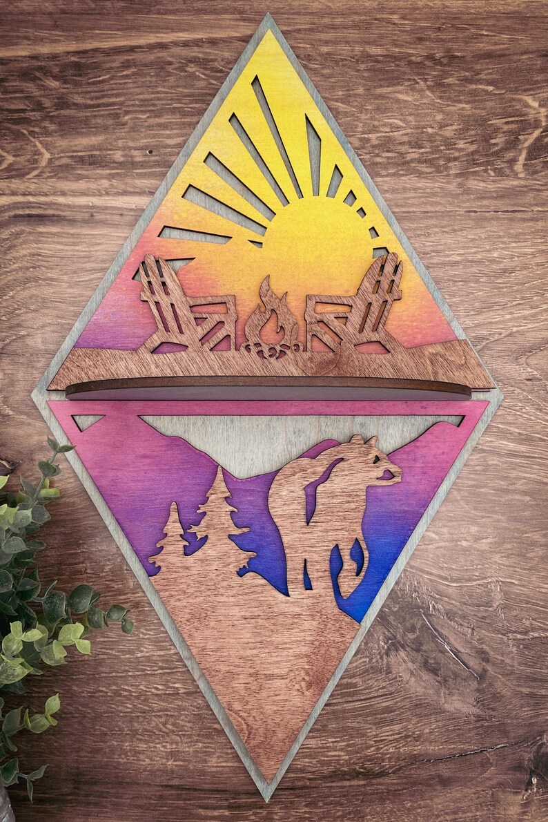 May include: A wooden diamond-shaped wall decor with a split design. The top half features a sunset scene with two chairs and a campfire. The bottom half features a mountain scene with a bear and two trees. The wood is stained in shades of yellow, orange, pink, purple, and blue.