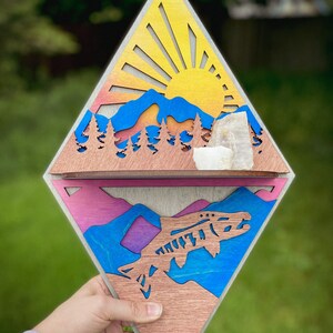 May include: A wooden diamond-shaped wall decor with a layered design. The top layer features a sunset scene with mountains, trees, and a sun with rays. The bottom layer features a blue and purple mountain scene with a fish jumping out of the water.