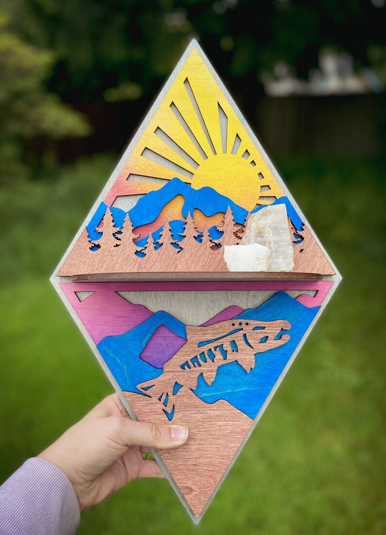 May include: A wooden diamond-shaped wall decor with a layered design. The top layer features a sunset scene with mountains, trees, and a sun with rays. The bottom layer features a blue and purple mountain scene with a fish jumping out of the water.