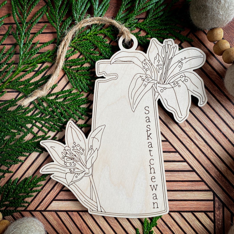 May include: Wooden ornament shaped like the province of Saskatchewan with a lily flower design and the text "Saskatchewan" written vertically on the right side.