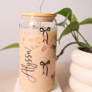 Può includere: Un bicchiere in vetro trasparente a forma di lattina riempito con una bevanda ghiacciata color beige, dotato di coperchio in bambù e cannuccia in vetro. L'esterno è decorato con fiocchi neri e il nome Alyssa in corsivo nero.
