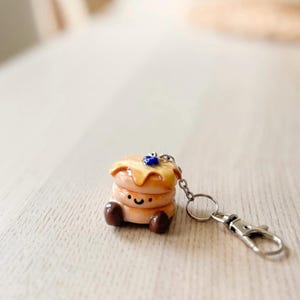 May include: A keyring featuring a miniature stack of two tan pancakes with a glossy yellow syrup glaze, a single dark blue berry on top, and a small smiling face drawn on the middle section, attached to a silver metal chain and lobster clasp.