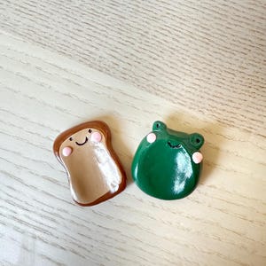 May include: Two handmade clay figurines: a slice of toast and a green frog. The toast is brown with a tan center and a smiling face. The frog is green with pink cheeks and a smiling face. Both are on a light wood surface.