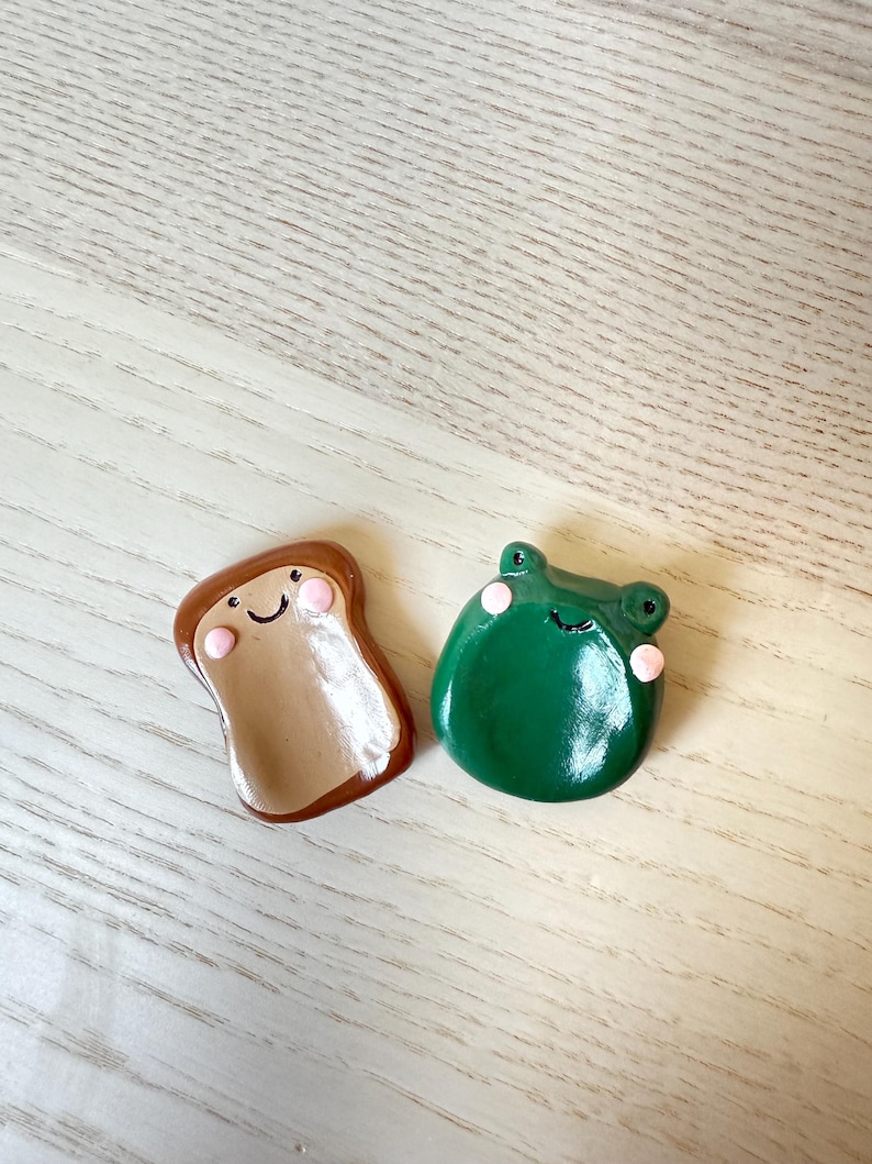 May include: Two handmade clay figurines: a slice of toast and a green frog. The toast is brown with a tan center and a smiling face. The frog is green with pink cheeks and a smiling face. Both are on a light wood surface.