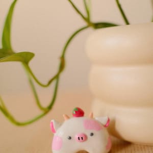May include: A small, white, ceramic cow figurine with pink spots, a pink snout, and two small horns. A tiny red strawberry with green leaves sits between the horns on top of the head. The cow has two dark eyes and pink inner ears.