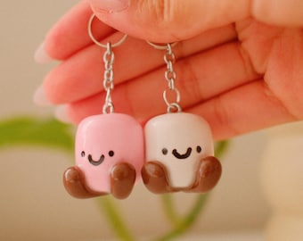 May include: Two keychains featuring miniature marshmallow figures, one light pink and one white. Each figure has a smiling face, small brown feet, and is attached to a silver-toned metal chain with a lobster clasp for secure attachment.