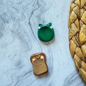 May include: Two handmade clay figurines: a green frog with pink cheeks and a slice of toast with a smiling face. The frog is round, and the toast is rectangular with a brown crust and a tan center. Both have a cute, cartoonish design.