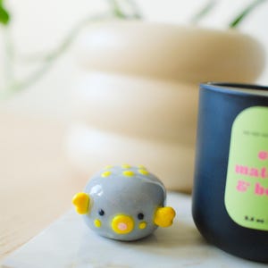 May include: A small, rounded gray ceramic pufferfish figurine featuring bright yellow fins, yellow spots on its back, and a central yellow mouth with a pink center. The figurine has two small black eyes and a smooth, glossy finish.