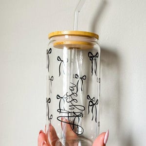 Pode incluir: Copo de vidro transparente com tampa de bambu e canudo de vidro. O copo é decorado com desenhos de laços pretos e a palavra "Hello" em fonte cursiva. Projetado para bebidas frias.