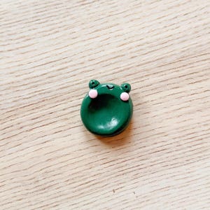 May include: A small, round, dark green frog-shaped object with pink cheeks and small, round eyes. The frog is sitting on a light brown wooden surface. The frog is a decorative item.